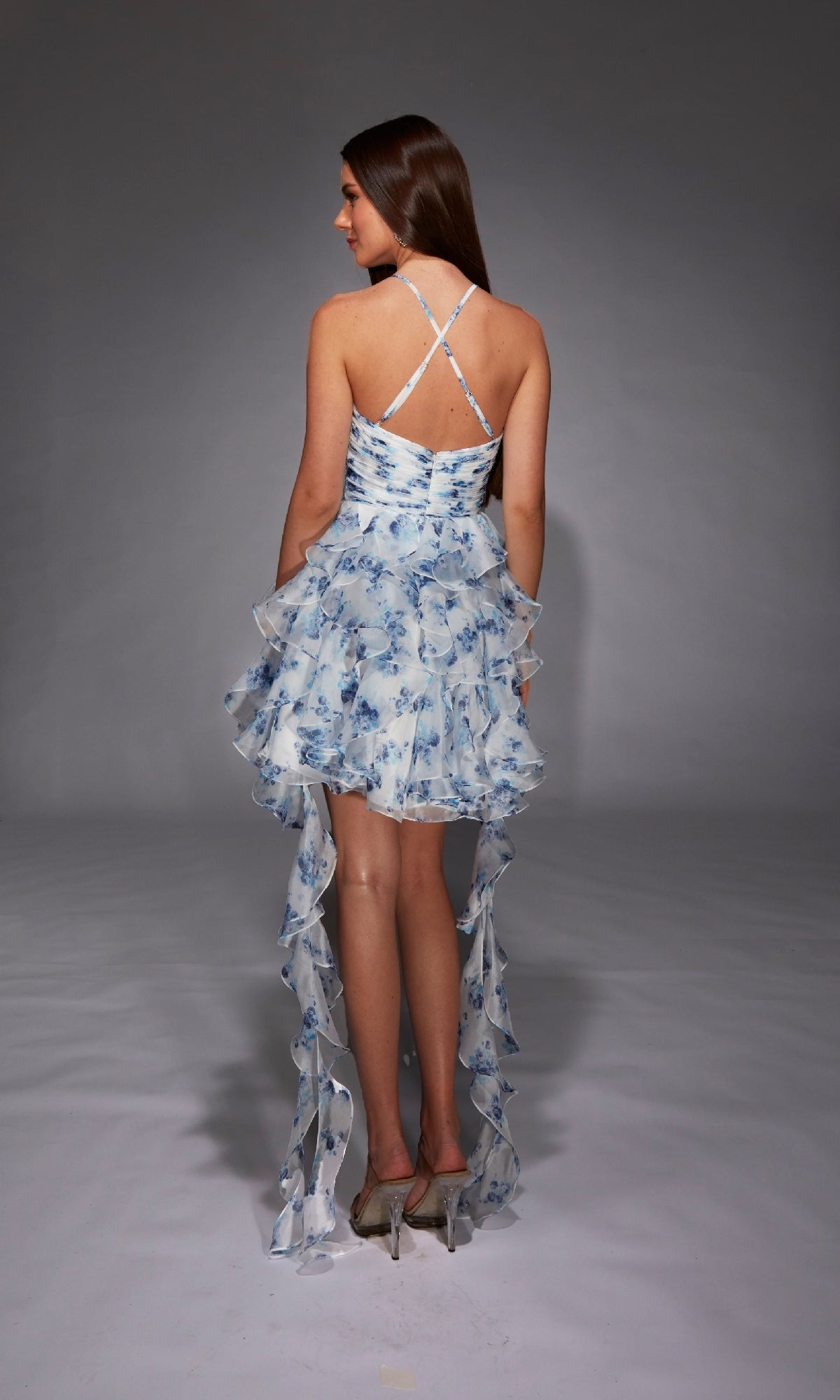 A woman stands in a studio, facing away, wearing the Alyce 30061 short homecoming dress—a white and blue floral high-low design with crisscross straps and an open back.